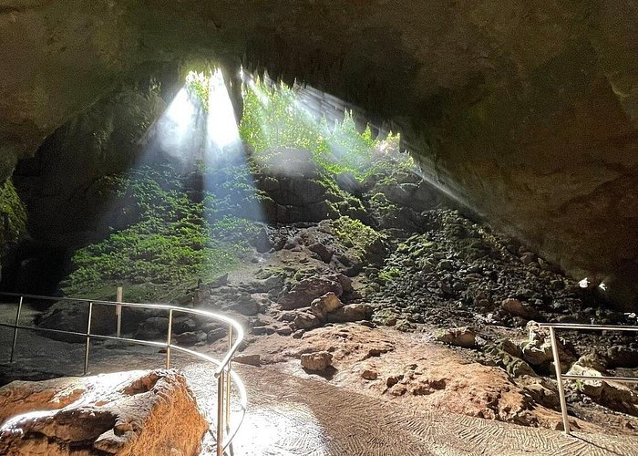 Sunlight entering a large cave in Belize with rocky pathways, natural stone formations, and lush greenery visible near the cave opening.