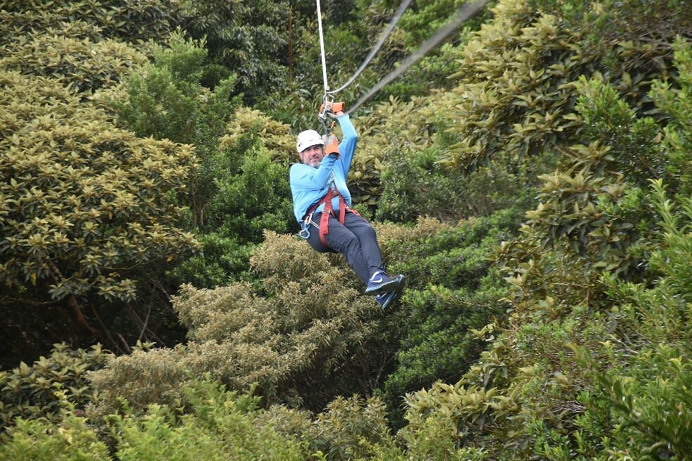 A zipliner wearing a helmet, blue long-sleeve top, dark pants, and a safety harness glides above dense green forest.