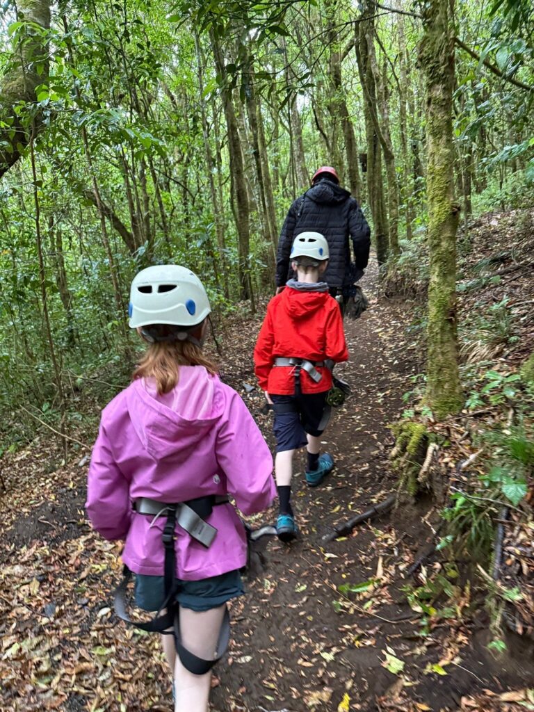 Two children wearing helmets and zipline harnesses follow a guide along a narrow trail through dense green forest.